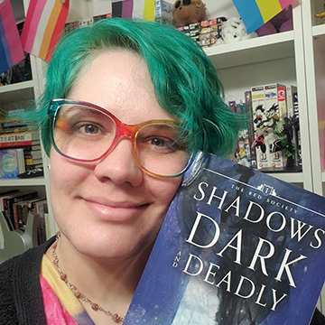 Andi Marie with teal hair and rainbow glasses smiles in front of a bookcase while holding up a copy of Shadows Dark and Deadly